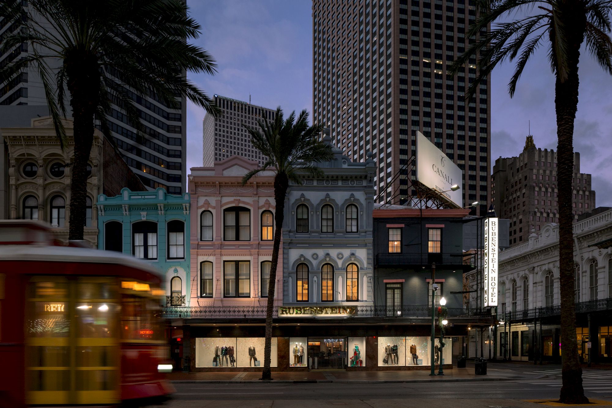 A street scene with colorful buildings, palm trees, and a passing red trolley car at dusk.