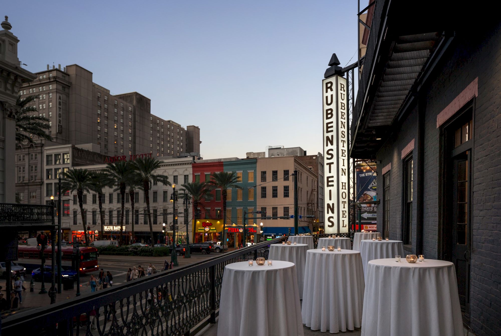 A city street scene at dusk with tall buildings, a "RUBENSTEIN'S" sign, palm trees, and tables set up on a balcony.