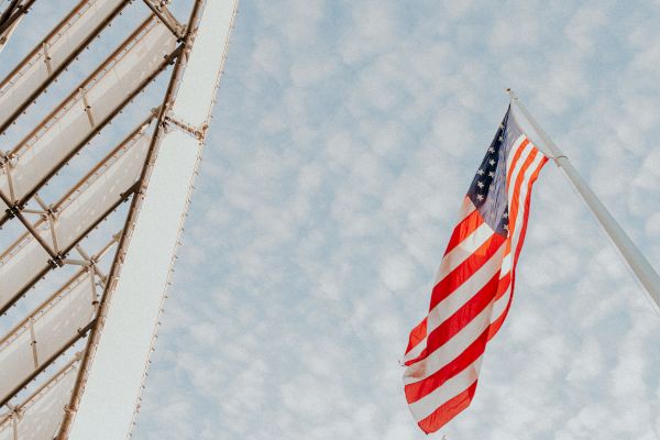 An American flag waves against a sky with scattered clouds, alongside part of a metal structure on the left.