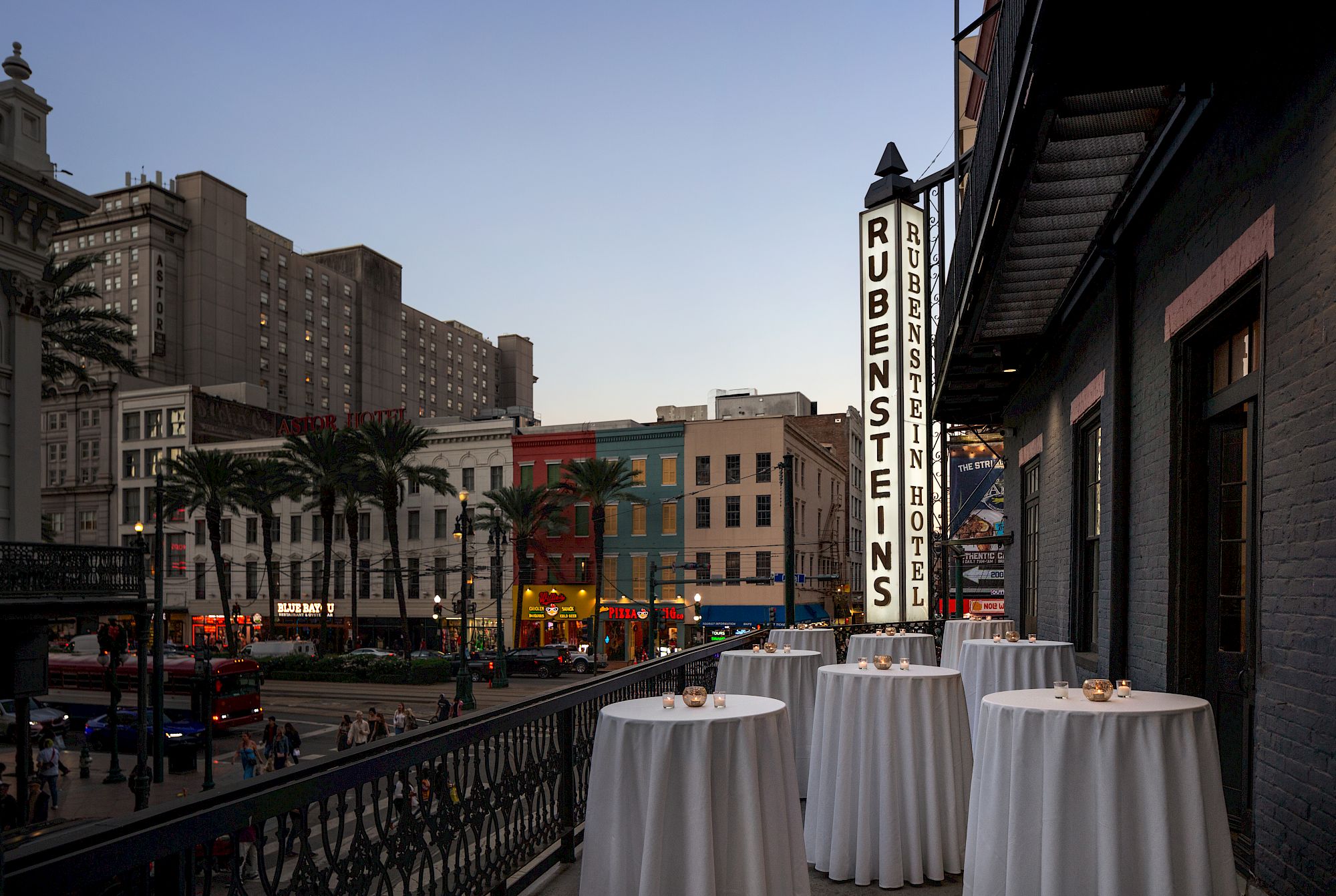 A city street scene at dusk features palm trees, tall buildings, and a sign for "Rubensteins" with tables set on a balcony.