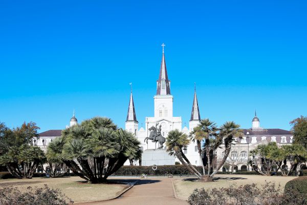 The image shows Jackson Square in New Orleans, featuring the iconic St. Louis Cathedral and an equestrian statue under a clear blue sky.