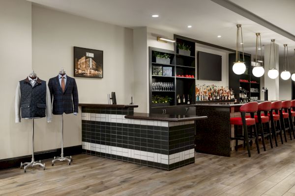 A stylish bar with red stools, mannequins in suits, modern lighting, and wooden flooring, creating a chic and inviting atmosphere.