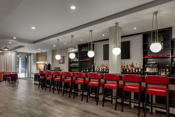 A modern bar with red stools, shelves of bottles, pendant lights, and a wood floor. Cozy seating area visible in the background.