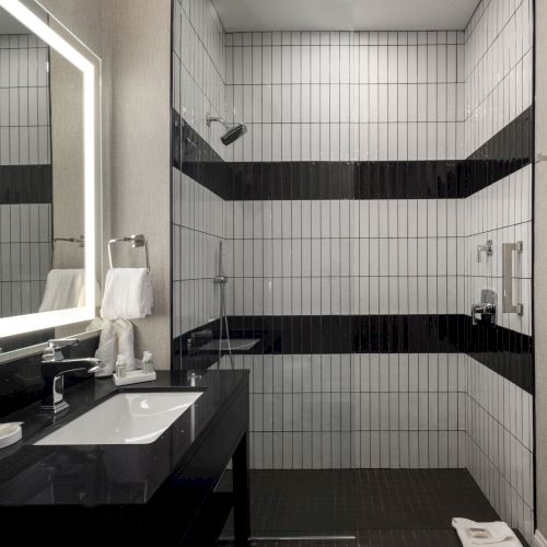 A modern bathroom with a black countertop, sink, lit mirror, and a walk-in shower featuring white tiles and black accents.