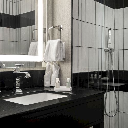 A modern bathroom with a large mirror, sink, black counter, towels, toiletries, and a walk-in shower with black and white tiles.