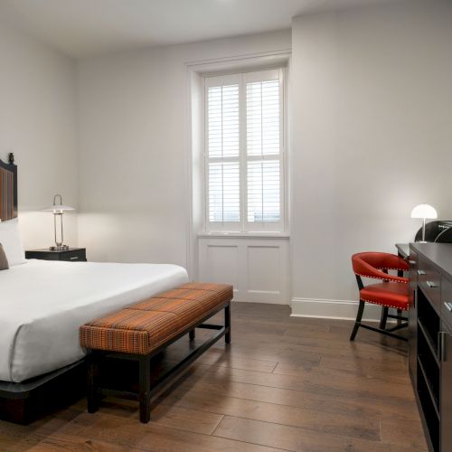 The image shows a modern hotel room with a bed, striped headboard, desk, chair, TV, and window with shutters, featuring wooden flooring.