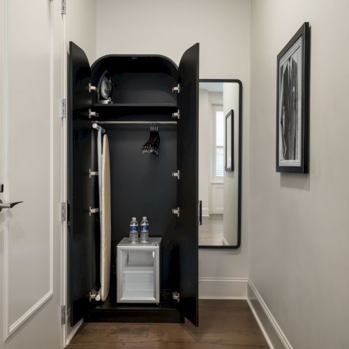 A hallway with an open closet containing two water bottles and hanging clothes; a mirror and framed picture are on the wall.