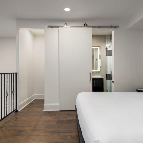 The image shows a minimalist bedroom with a bed, sliding barn door, and modern railing, leading to a small bathroom.