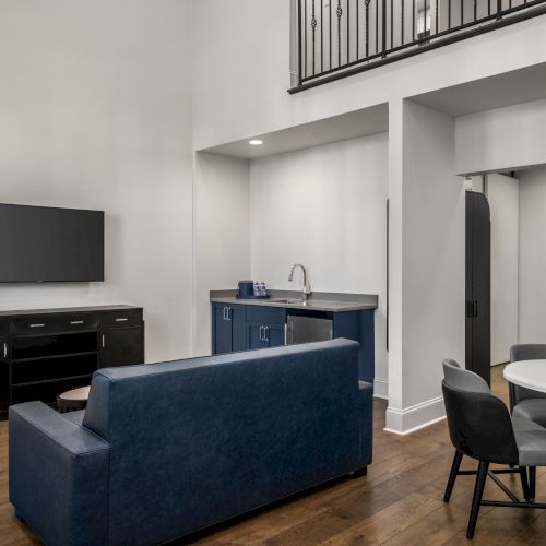 A modern living space with a sofa, TV, kitchenette, round dining table, and artwork on the wall. Warm lighting and wood flooring.