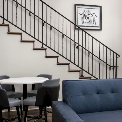 The image shows a modern interior with a staircase, a round table with four chairs, a blue sofa, and a framed picture on the wall.