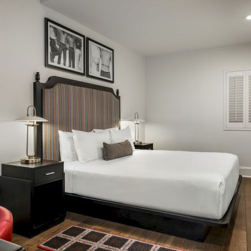 A modern hotel room with a bed, two lamps, wall art, a TV, and a small desk with a red chair, all in neutral tones and wood flooring.