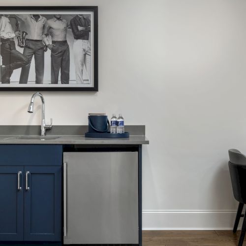 A kitchenette features navy cabinets, a sink, a mini-fridge, and a framed photo. Adjacent is a black chair and a small table.