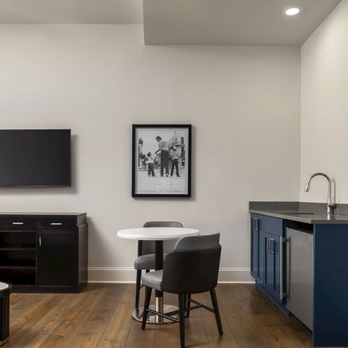 The image shows a modern room with a TV, a small round table with chairs, a cabinet with a sink, and a black and white photo on the wall.