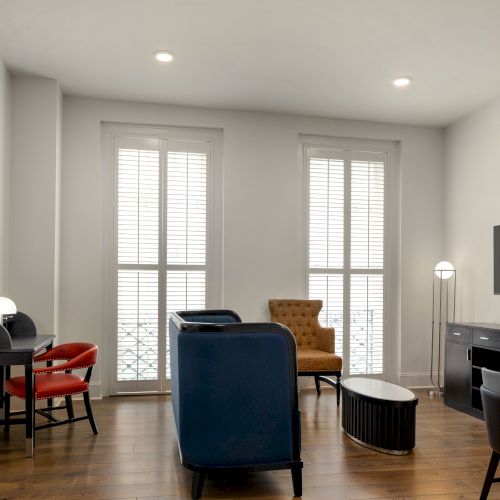 A modern living room with assorted chairs, a desk, and a wall-mounted TV, featuring wooden floors and large windows with blinds.