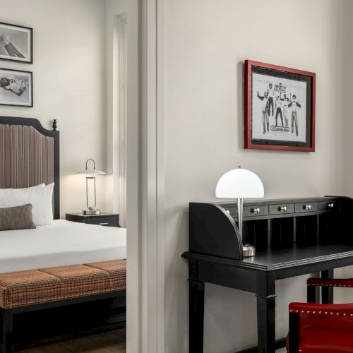 The image shows a stylish hotel room with a bed, artwork, a desk, lamp, and red chairs, creating a modern and elegant atmosphere.