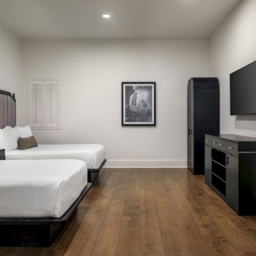 The image shows a modern hotel room with two beds, a TV, framed wall art, and a sleek desk. The decor is minimalist with wooden flooring.