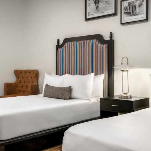 The image shows a hotel room with two beds, a nightstand with a lamp, chairs, and wall art, all in a modern, neutral-toned decor.