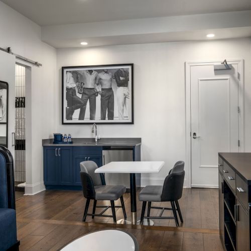 A modern room with a small table and chairs, a wall-mounted TV, and a framed photo. Dark cabinets and a door are visible.