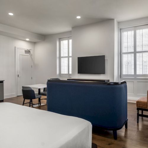 A modern room with a bed, blue sofa, TV on the wall, small dining area, and wicker chair, featuring a large window with shutters.