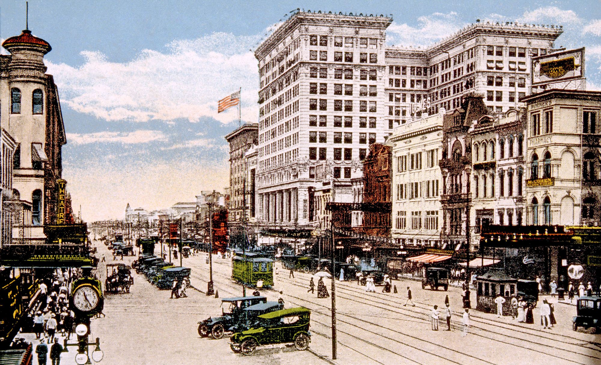 A vintage city street scene with classic cars, pedestrians, and historic buildings under a blue sky with an American flag visible.