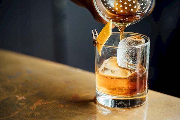 A cocktail being poured into a glass with a large ice cube and garnished with a twist of citrus peel on a bar counter.