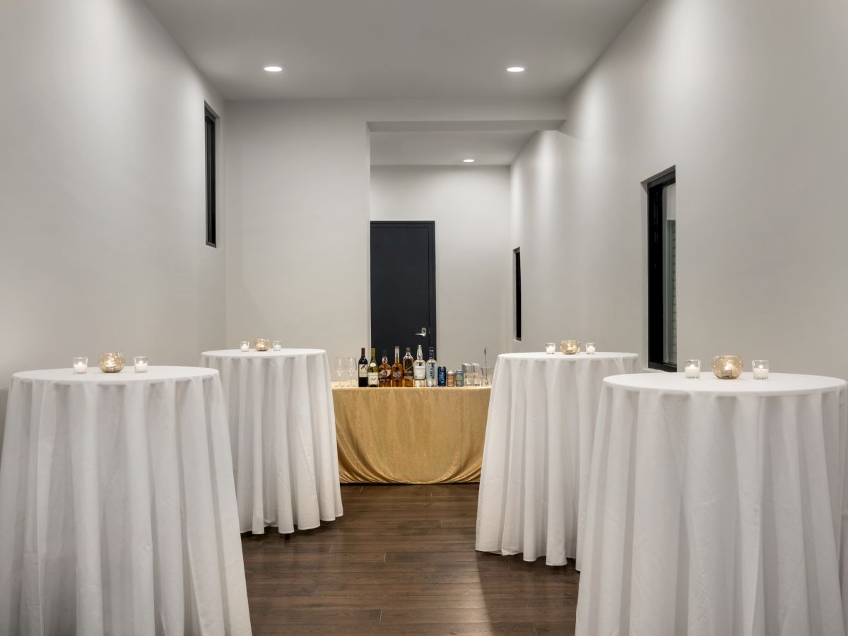 The image shows a narrow room with cocktail tables covered in white tablecloths and a table with drinks against the back wall.