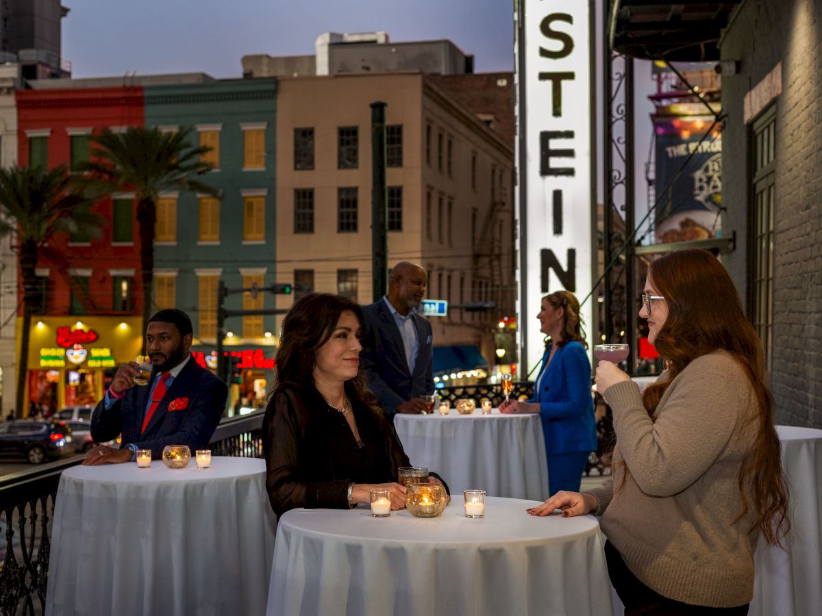 People conversing at outdoor tables adorned with candles on a balcony, with colorful buildings and a "STEIN" sign in the background.