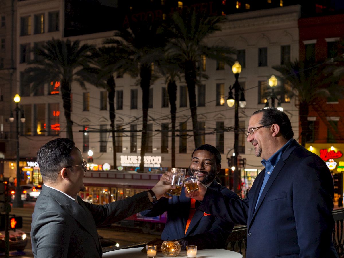 Three people stand around tables with candles, having a conversation outdoors at night in a city setting.