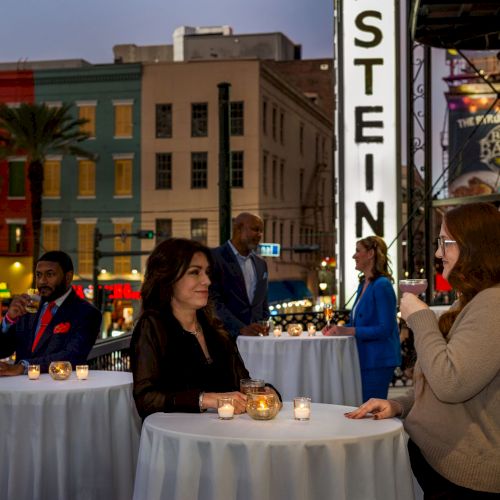 People are socializing around tables with candles outside, near brightly colored buildings and a sign reading "STEIN".