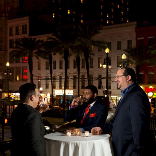 Three people are standing around a cocktail table outdoors at night, enjoying drinks, with city lights and buildings in the background.