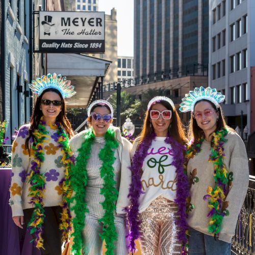 Four people wearing festive Mardi Gras outfits with feather boas and headpieces stand on a city street.