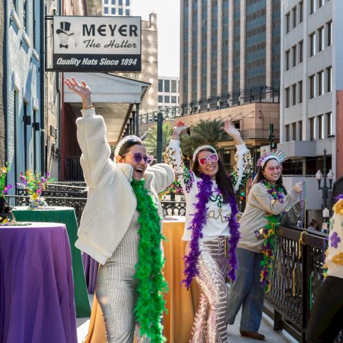 People celebrating on a balcony with colorful attire and beads, featuring a sign for Meyer the Hatter in the background.