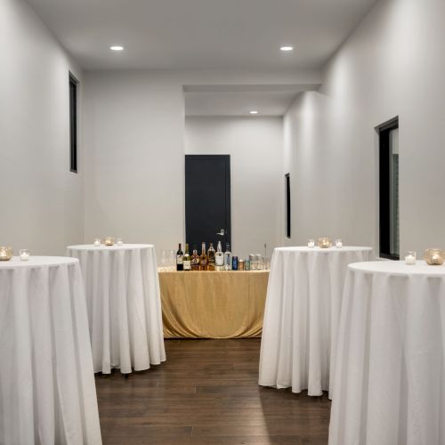 The image shows a narrow room with four round tables covered in white cloth and a table at the far end with bottles and a beige tablecloth.