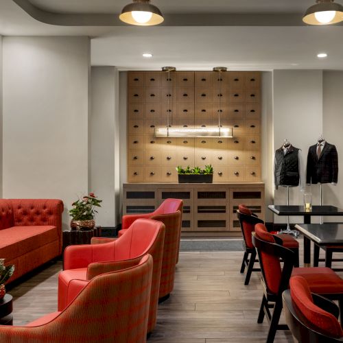 A modern lounge with red seating, tables, suits on display, plants, and soft lighting, creating a stylish and cozy ambiance.