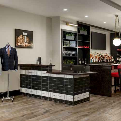 The image shows a modern bar with red chairs, mannequins dressed in suits, a tiled counter, and shelves with glasses and bottles.