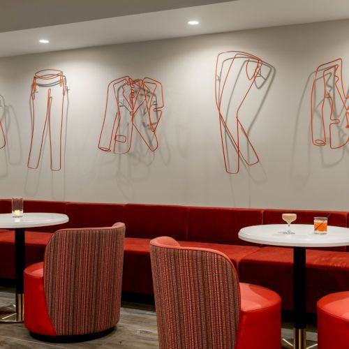 The image shows a modern lounge with red seating, white tables, and wire art on the wall resembling clothing items.