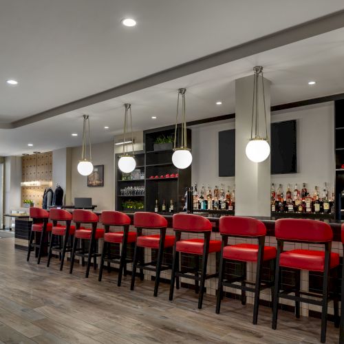 A modern bar with red chairs, hanging lights, and a well-stocked counter. The space is open and inviting, ready for guests.